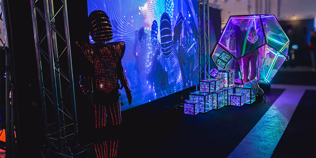 LED-klädd performer vid stor skärm; till höger en neonbelyst installation med genomskinlig polyeder och staplade ljuskuber, med besökare inuti, på mässgolvet.