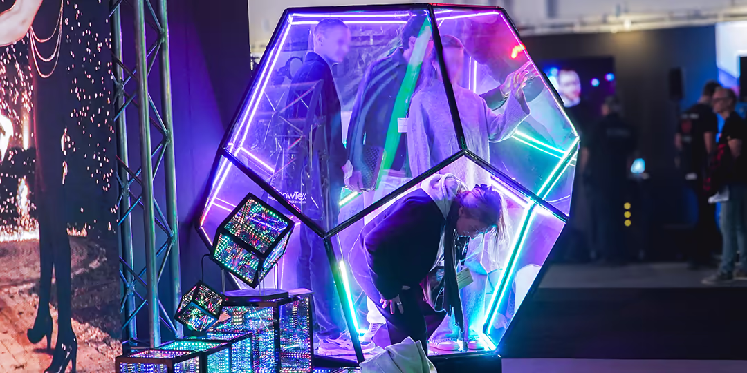 Interaktiv ljusinstallation på mässa: genomskinlig, neonbelyst polyeder med besökare inuti och staplade LED-kuber intill.